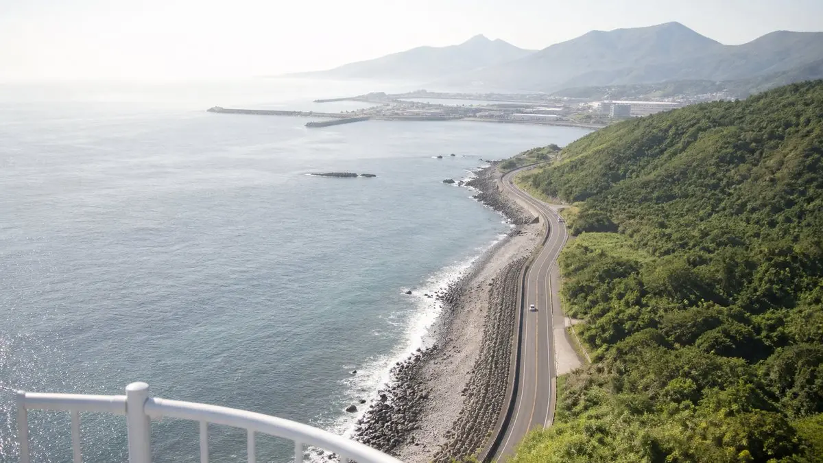 從燈塔塔頂俯瞰整段海岸線，公路沿著海岸蜿蜒，遠處是港口與山